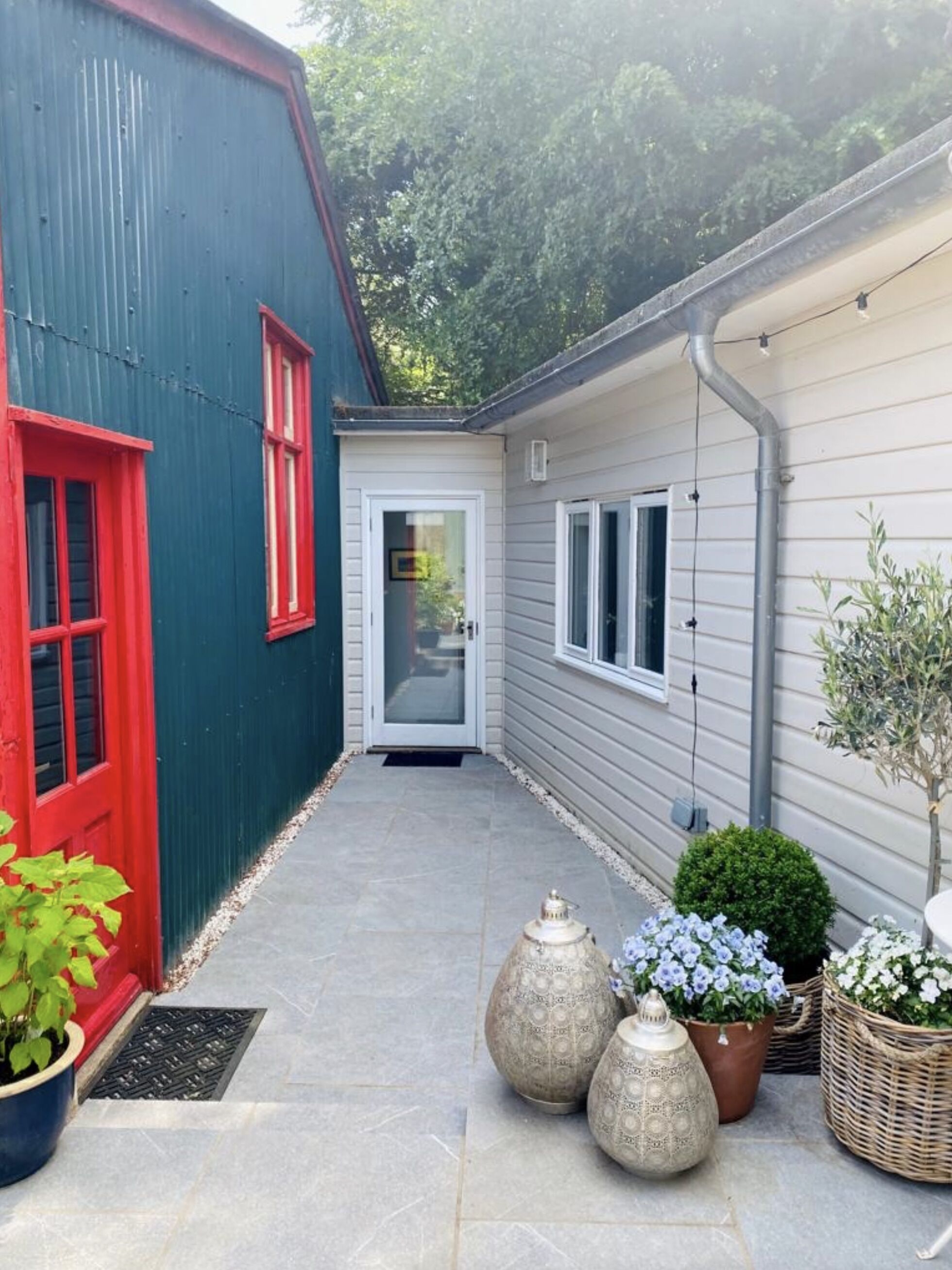 Un étroit sentier mène à une maison avec une porte rouge et une porte blanche, flanquée d’un bâtiment bleu et d’une maison grise. Des plantes en pot et un petit arbre se trouvent le long du sentier.