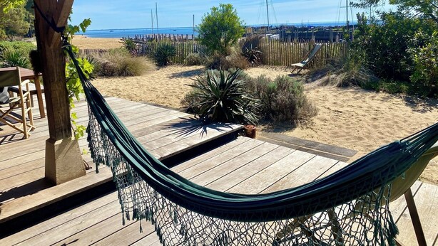 Un hamac est suspendu sur une terrasse en bois, d’où l’on a vue sur une plage, une clôture en bois et quelques plantes.