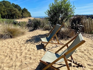 Two wooden lounge chairs on a sandy beach with a wooden fence and vegetation in the background.