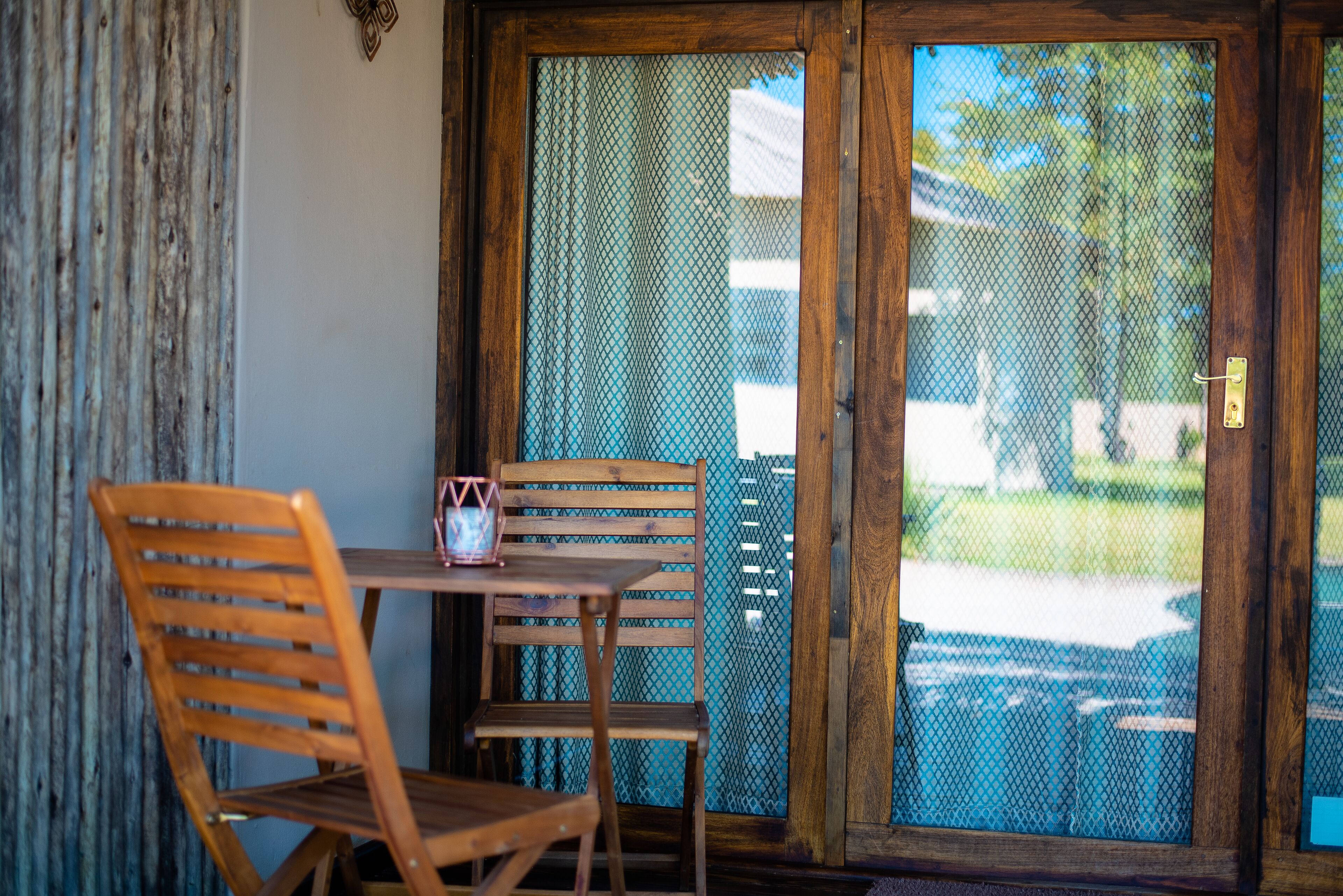 A wooden chair and table near a glass door with a metal screen, leading to an outdoor area with a pool.