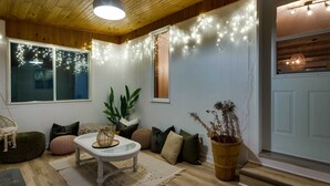 Ein gemütliches Wohnzimmer mit Holzbalkendecke, einem weißen Tisch mit Kissen, einer Topfpflanze und Lichterketten.