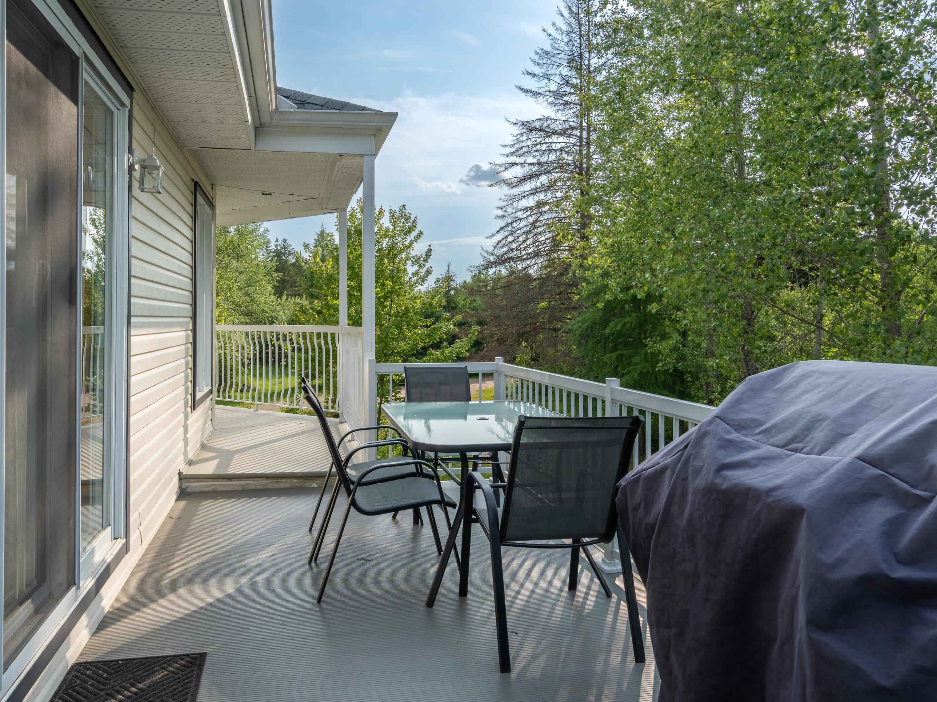 Une terrasse extérieure couverte, avec une table et des chaises, un barbecue et une vue sur des arbres et une maison au loin.