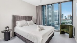 A modern hotel room with a large bed, a round side table, a green armchair, and a city view through large windows. - Luxury Signature Stay Ocean Views 2BR Lvl 9 (Broadbeach)