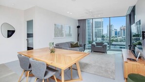 A modern living room with a wooden dining table, grey chairs, a grey sofa, and a large window offering a city view. - Luxury Signature Stay Ocean Views 2BR Lvl 9 (Broadbeach)
