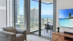 A modern living room with a grey sofa, a wooden TV stand, and a large TV displaying a beach scene. - Luxury Signature Stay Ocean Views 2BR Lvl 9 (Broadbeach)