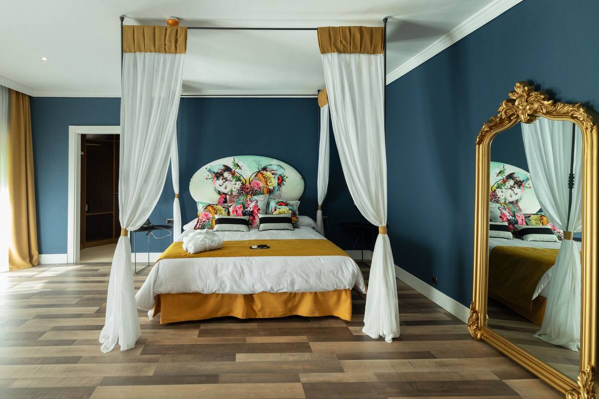 A four-poster canopy bed with a floral headboard, white drapes, and a gold bed skirt.