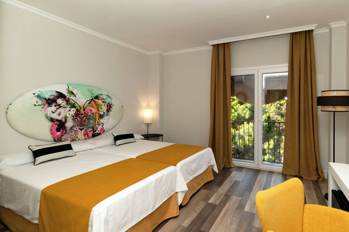 A hotel room with a large bed, a yellow armchair, a painting on the wall, and a view of greenery outside.