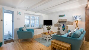 A living room with a sofa, a coffee table, and a TV. There is a window with blinds, a door, and a stone wall with a painting. - 20 The Warren, St Ives – Sleeps 4 – Outside Space (St Ives)