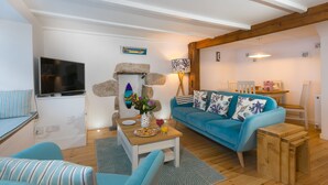 A cozy living room with a blue sofa, a wooden coffee table, and a TV. There is a stone wall decoration and a lamp on a side table. - 20 The Warren, St Ives – Sleeps 4 – Outside Space (St Ives)