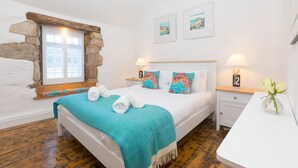 A bedroom with a stone-framed window, a bed with a teal blanket, two pillows, a nightstand with a lamp, and a vase with flowers. - 20 The Warren, St Ives – Sleeps 4 – Outside Space (St Ives)