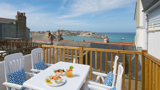 A terrace with a table set for two, overlooking a coastal town with historic buildings and a harbor. - 20 The Warren, St Ives – Sleeps 4 – Outside Space (St Ives)