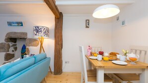 A cozy dining area with a wooden table set for breakfast, a blue sofa, and a lamp with a patterned shade. - 20 The Warren, St Ives – Sleeps 4 – Outside Space (St Ives)