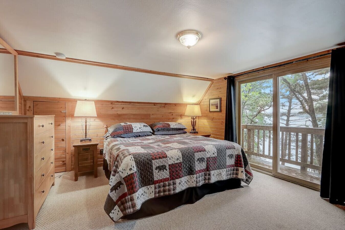 Un dormitorio con cabecera de madera, cama con colcha, mesita de noche con lámpara, cómoda y ventana con vista a árboles.