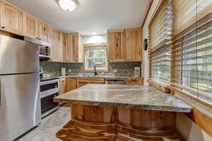 A kitchen with wooden cabinets, a stainless steel refrigerator, a granite countertop, and a window with blinds.