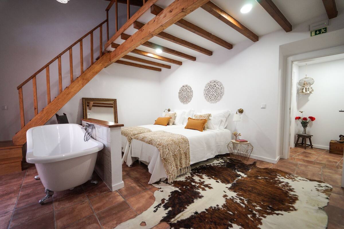 A bedroom with a wooden staircase, a white bathtub, a bed with white bedding and a cowhide rug.
