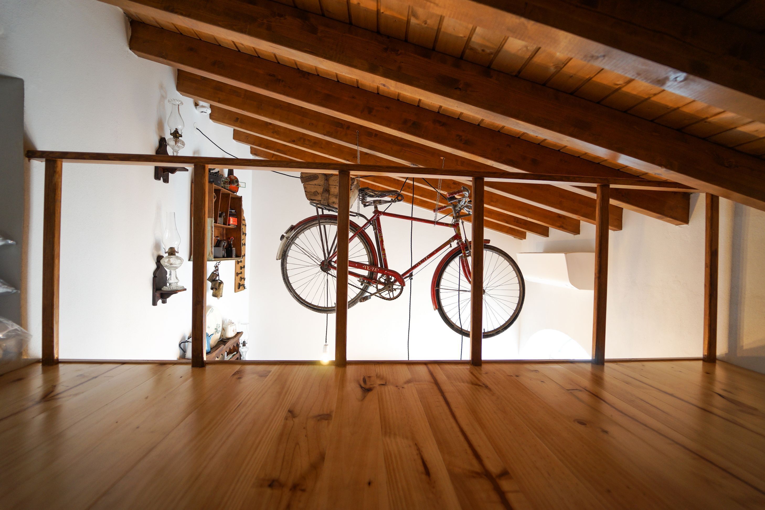 A room with wooden flooring, a red bicycle hanging from a wooden beam, and a white wall with a shelf containing various items.