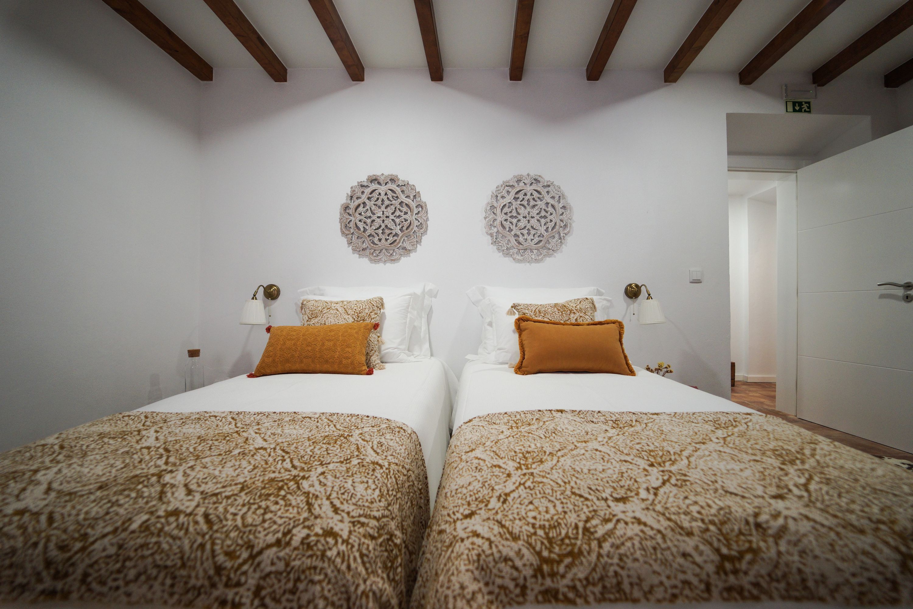A hotel room with two beds, each with patterned bedding and a single orange pillow. There are decorative wall hangings above the beds and a door to the right.