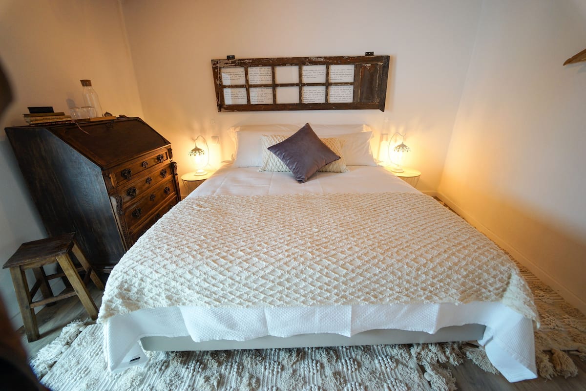 A neatly made bed with a quilted cover, a wooden headboard with a plaque, bedside lamps, and a small wooden stool.