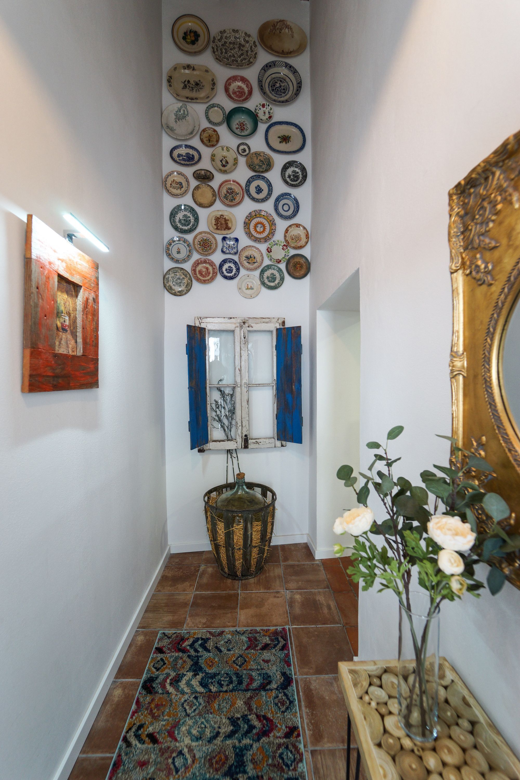 Un pasillo con un tapete estampado, una pared decorada con platos, una ventana con contraventanas azules y un jarrón con flores.