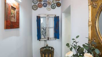 Un pasillo con un tapete estampado, una pared decorada con platos, una ventana con contraventanas azules y un jarrón con flores.