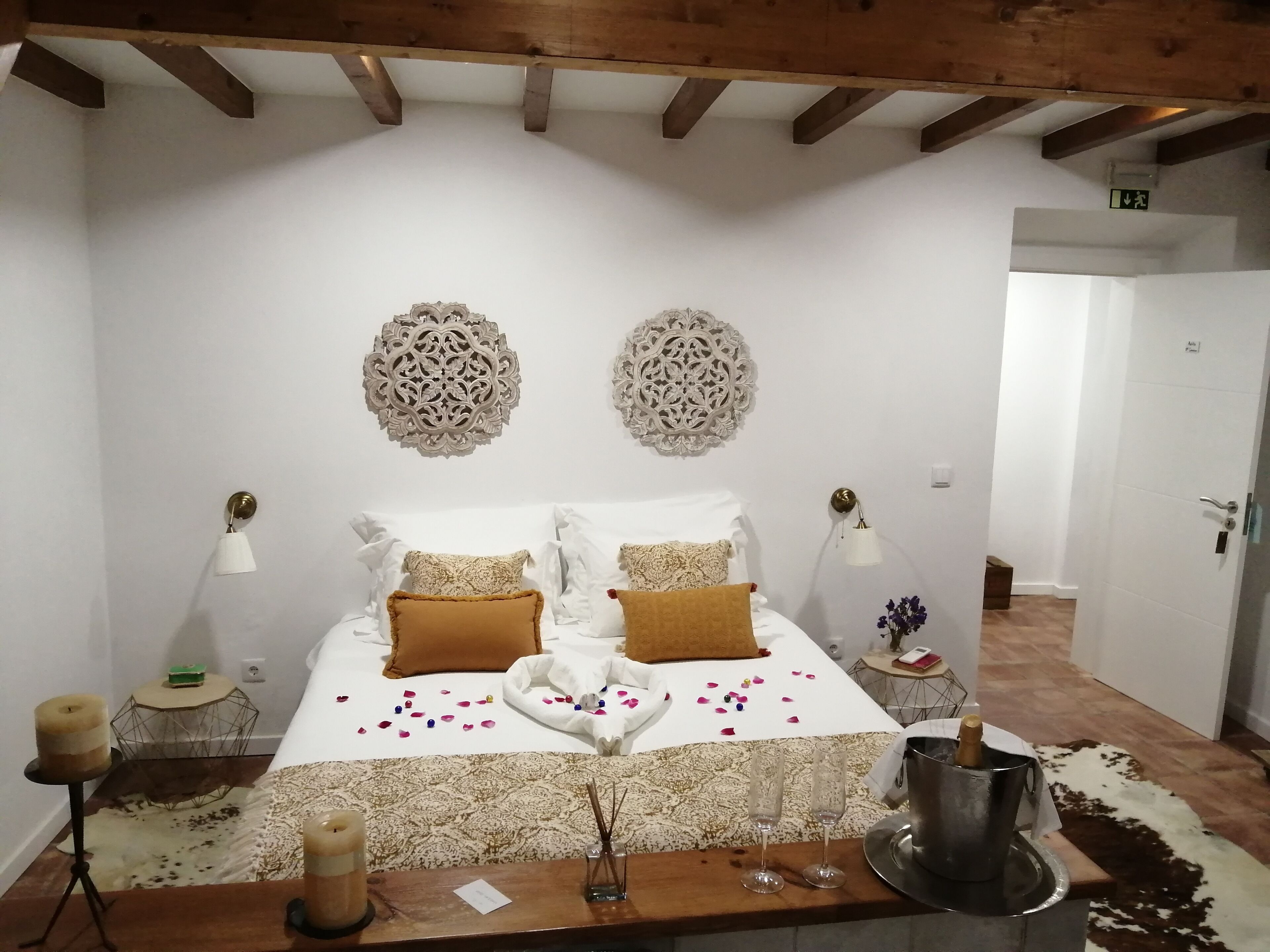 A hotel room with a bed, decorative wall pieces, a bedside table with a candle, a small round table with a green object, and a hallway with a door.