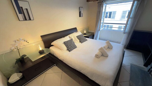 Een keurig opgemaakt bed met witte lakens, twee gestreepte kussens en opgerolde handdoeken op een donker houten bedframe. Een klein tafeltje met een lamp en een bloempot staat naast het bed.