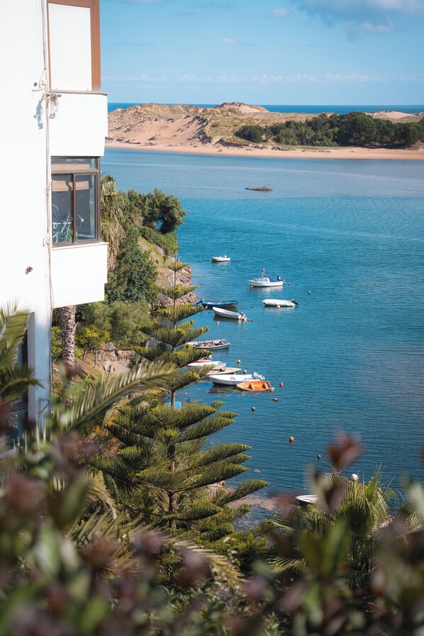 A coastal view with boats on the water, a sandy beach, and a building with a balcony overlooking the sea.