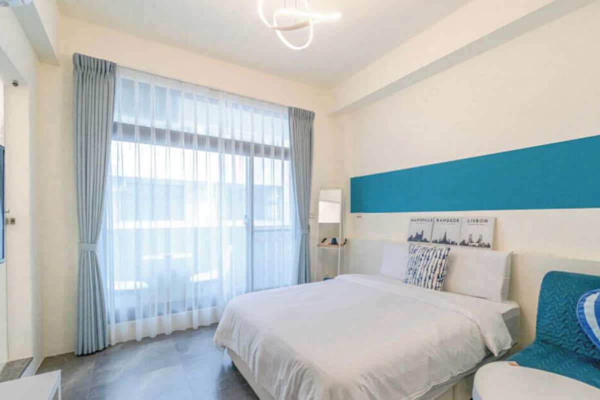 A hotel room with a large bed, a bedside table, a nightstand, a window with curtains, and a wall-mounted light fixture.
