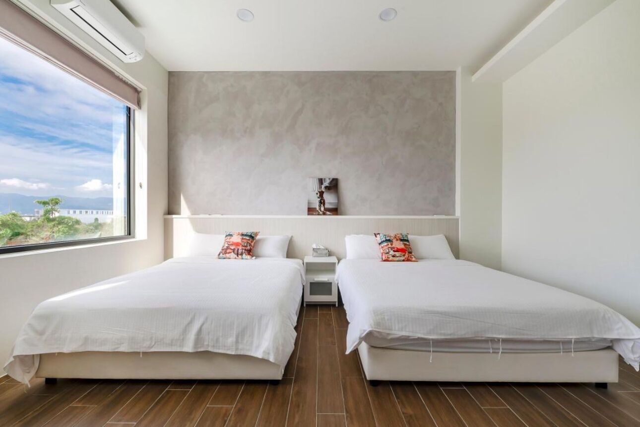 A modern hotel room with two single beds, a large window with a view, a bedside table with a lamp, and a neutral-colored wall.