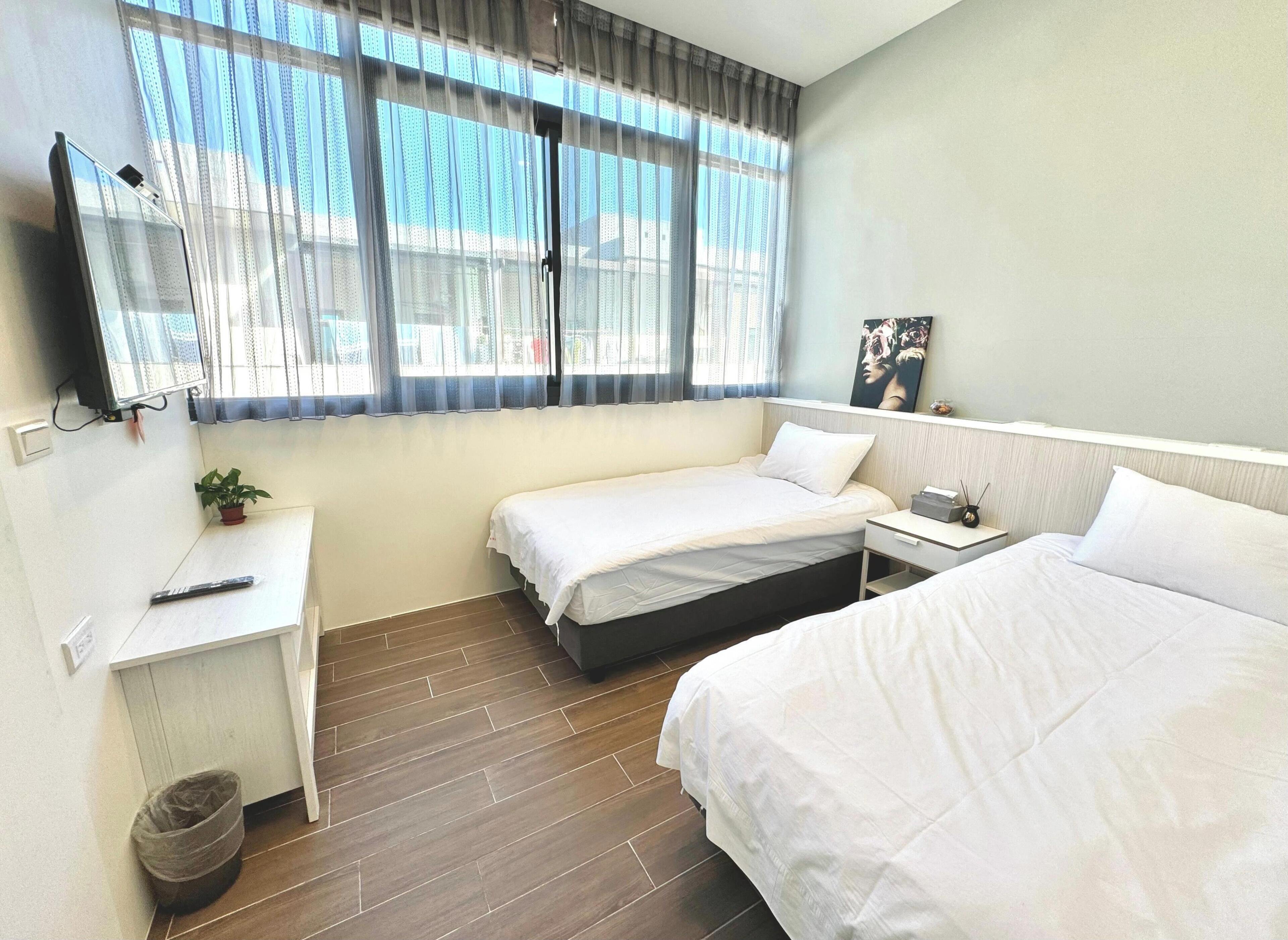 Ein Hotelzimmer mit zwei Betten, einem Schreibtisch, einem an der Wand montierten Fernseher und einem Fenster mit leichtem Vorhang.