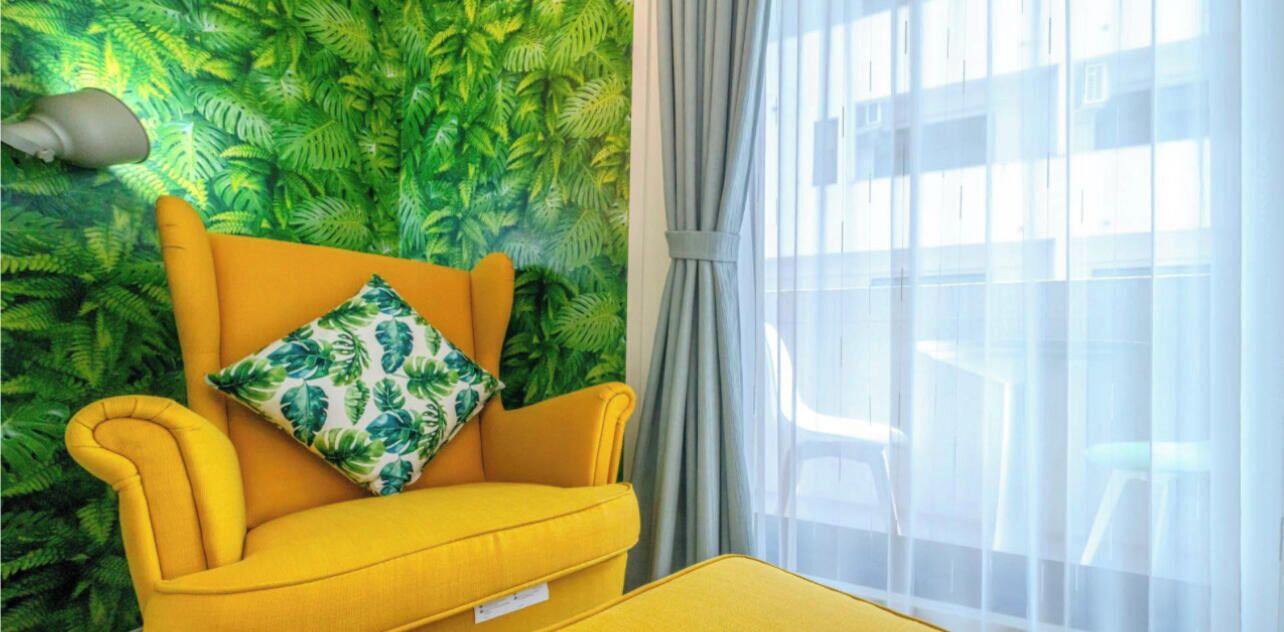Un fauteuil jaune avec un coussin orné d’un motif de feuilles, adossé à un mur vert couvert de verdure, une fenêtre avec des rideaux légers et une table jaune.