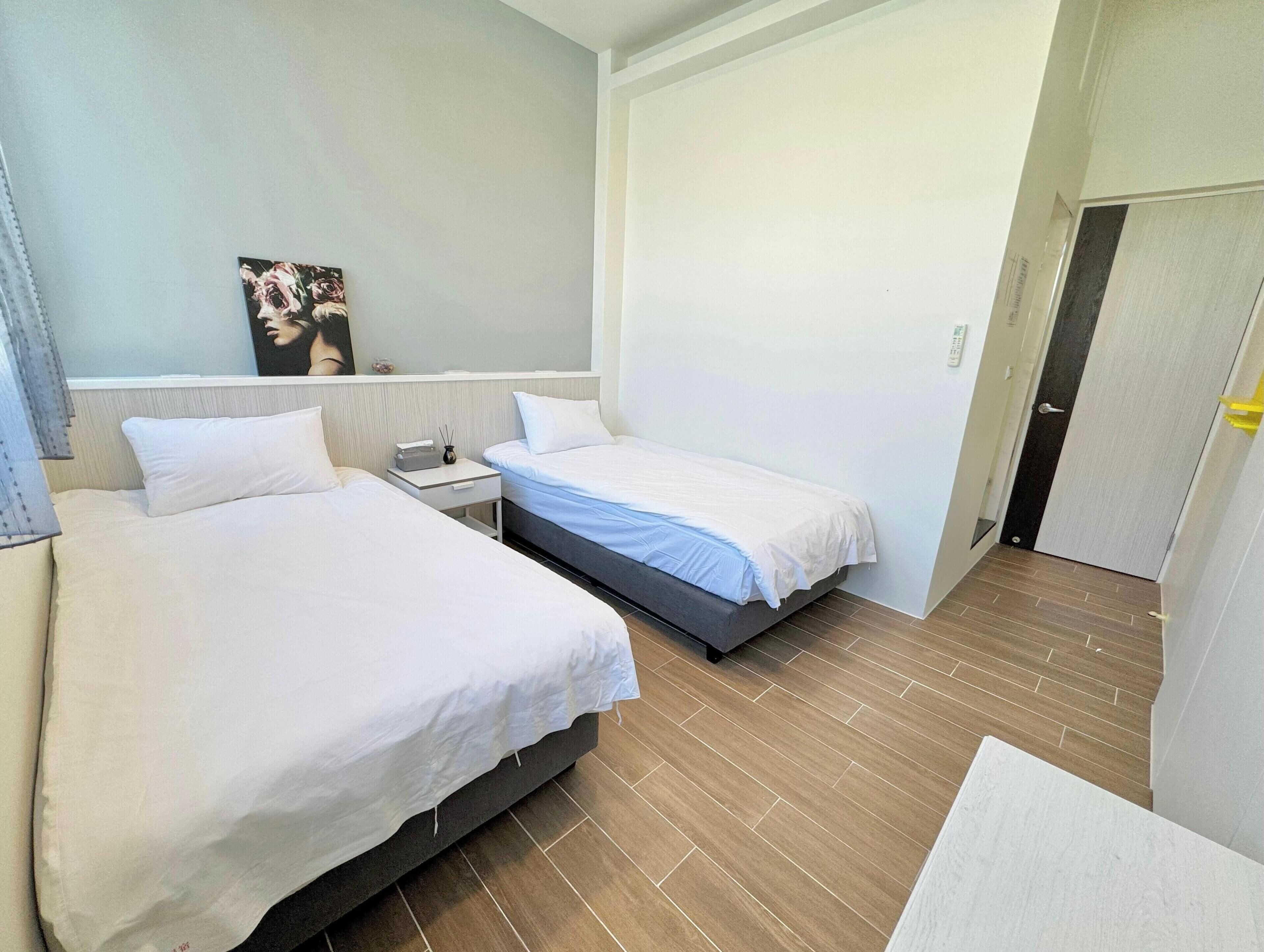 Ein Hotelzimmer mit zwei Betten, einem kleinen Nachttisch mit Lampe, einem Bild an der Wand und einer Tür rechts.