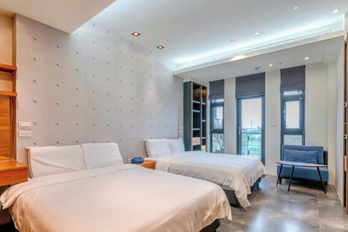 A hotel room with two beds, a desk, a chair, a TV, and a view of the city through large windows.