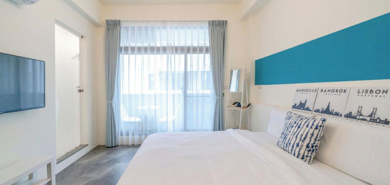 A hotel room with a large bed, a flat-screen TV, a window with sheer curtains, and a wall with cityscape artwork.