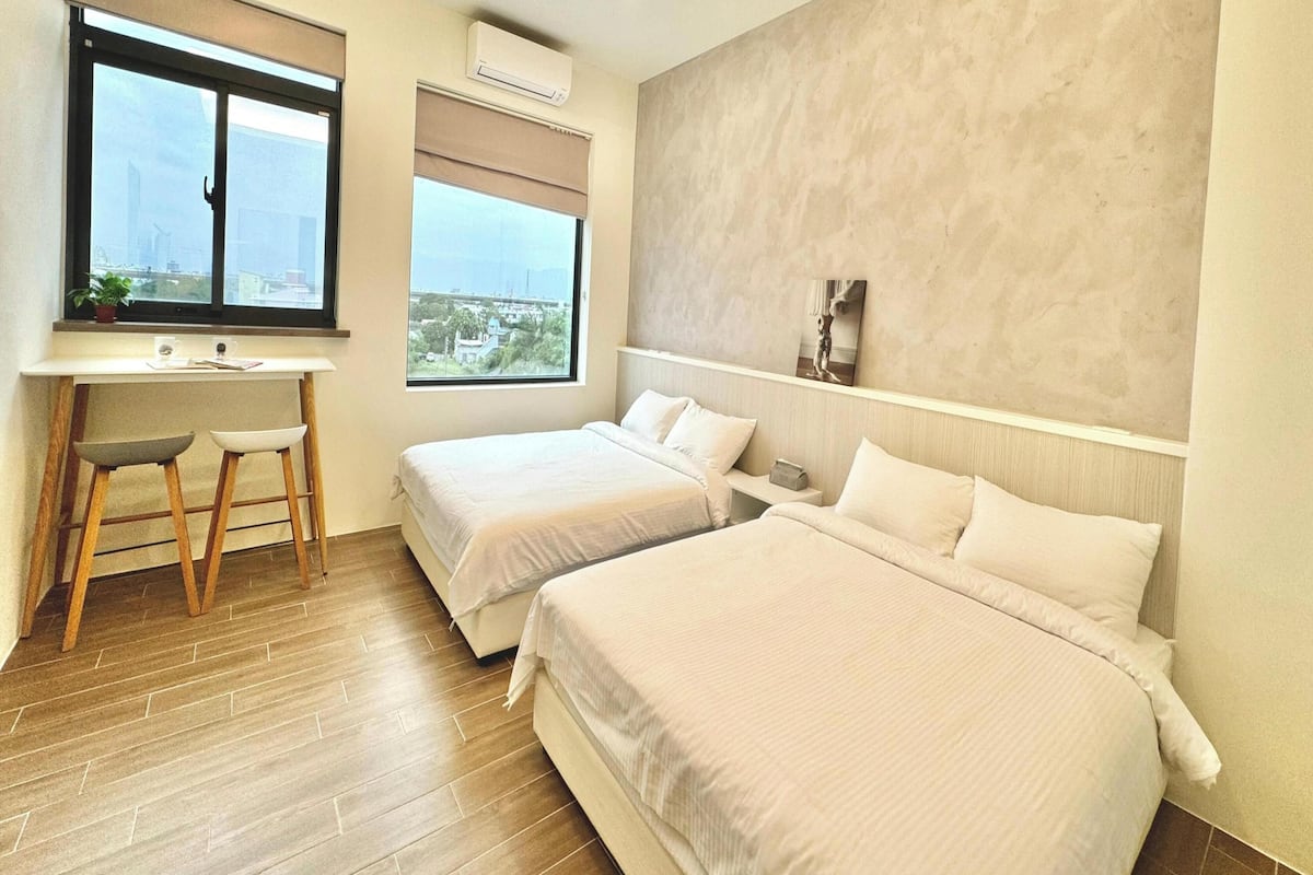 A hotel room with two beds, a desk with a chair, a window with a view, and an air conditioning unit.