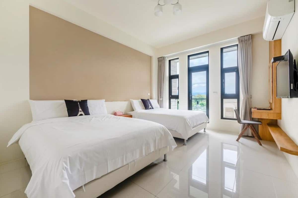 A hotel room with two beds, a desk, a TV, and large windows.