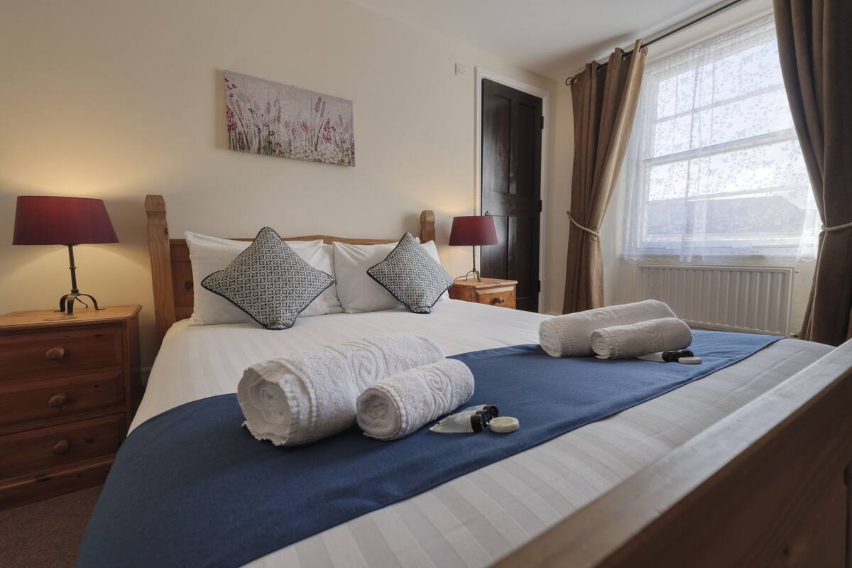A neatly made bed with rolled towels, a blue bedspread, and a wooden headboard. There are two bedside tables with lamps and a painting on the wall.