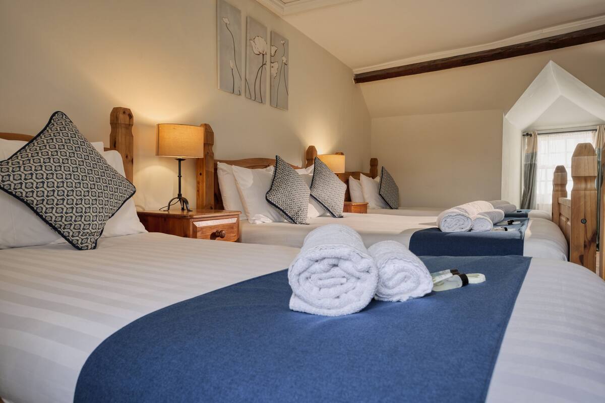 A hotel room with two beds, each with a blue bedspread and white linens. There are patterned pillows, a bedside table with a lamp, and a window with curtains.