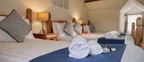 Habitación de hotel con dos camas, cada una con colcha azul y ropa de cama blanca. Hay almohadas estampadas, una mesita de noche con lámpara y una ventana con cortinas.
