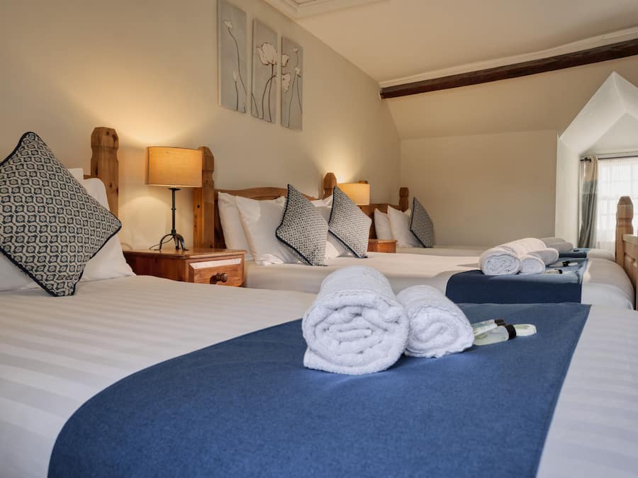 Een hotelkamer met twee bedden, elk met een blauwe sprei en witte lakens. Er zijn kussens met een patroon, een nachtkastje met een lamp, en een raam met gordijnen.