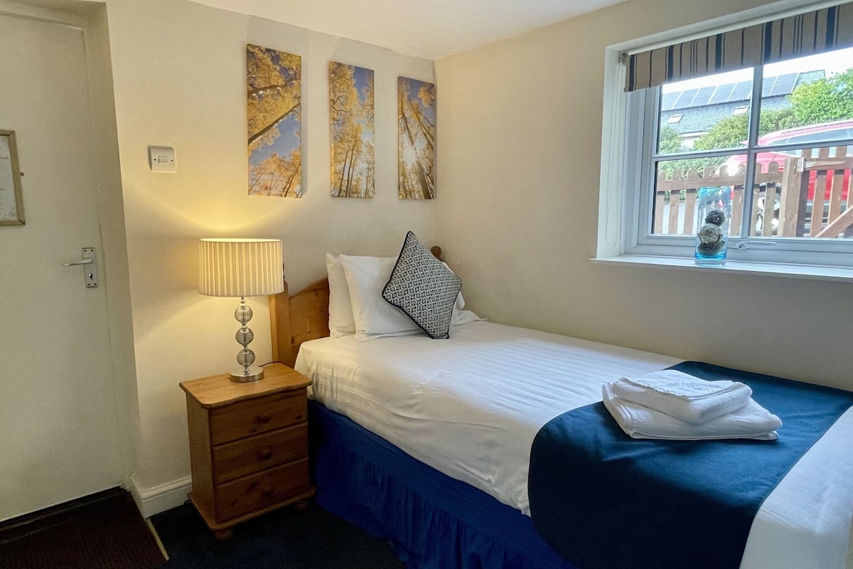 A bedroom with a bed, a nightstand, a lamp, and a window with a view of the outside.