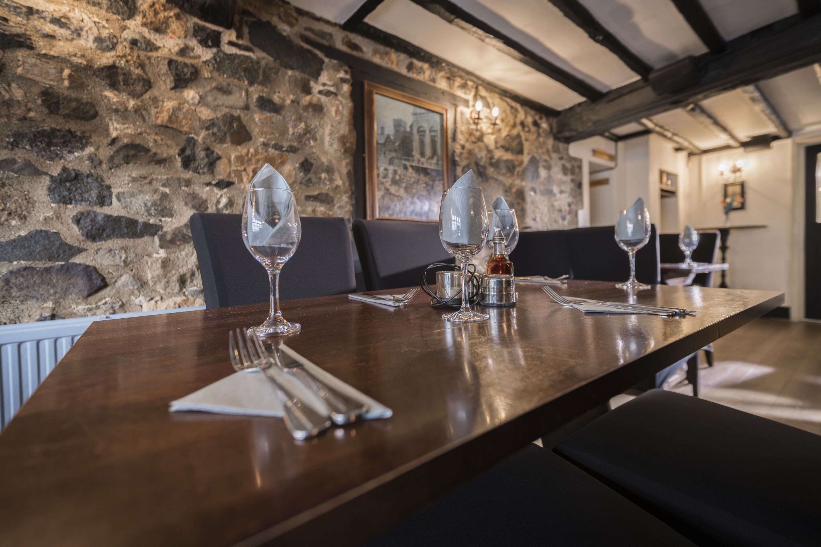 Una mesa de comedor dispuesta con copas de vino, una jofaina y cubiertos en una habitación con paredes de piedra y vigas de madera.
