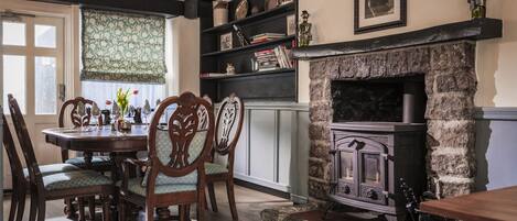 A dining area with a round wooden table, four upholstered chairs, a stone fireplace, and a bookshelf with various items.