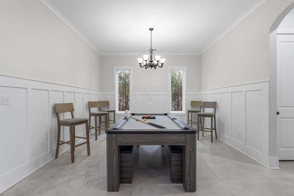 Une table de billard dans une pièce aux murs blancs et au lambris en bois, entourée de quatre chaises.