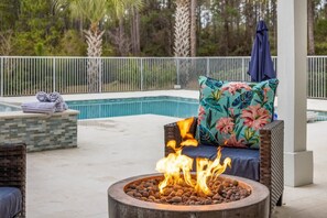 A poolside area with a fire pit, wicker furniture, and a view of the pool and surrounding trees.