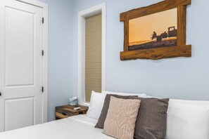 A bedroom with a bed, pillows, a nightstand, a framed picture on the wall, a door, and a window with blinds.