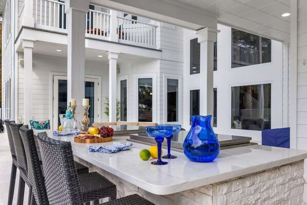 A spacious outdoor dining area with a white marble counter, wicker chairs, and a variety of table settings including a fruit bowl and blue glassware.