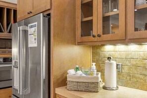 A kitchen with wooden cabinets, a stainless steel refrigerator, a basket with toiletries, and a paper towel dispenser.