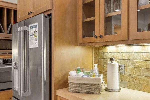 A kitchen with wooden cabinets, a stainless steel refrigerator, a basket with toiletries, and a paper towel dispenser.