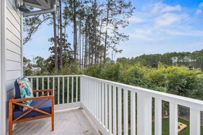 Un balcón con baranda blanca, una silla de madera con cojín y vistas a árboles y vegetación.